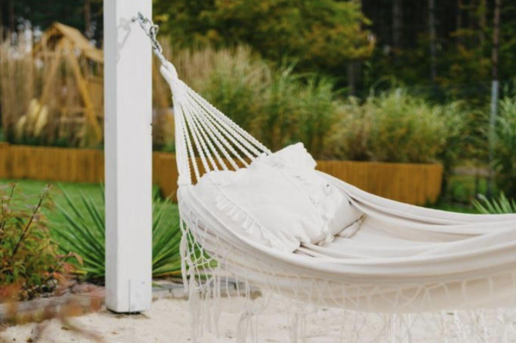 A hammock tied to a white post, set in a garden with greenery and a wooden fence in the background.