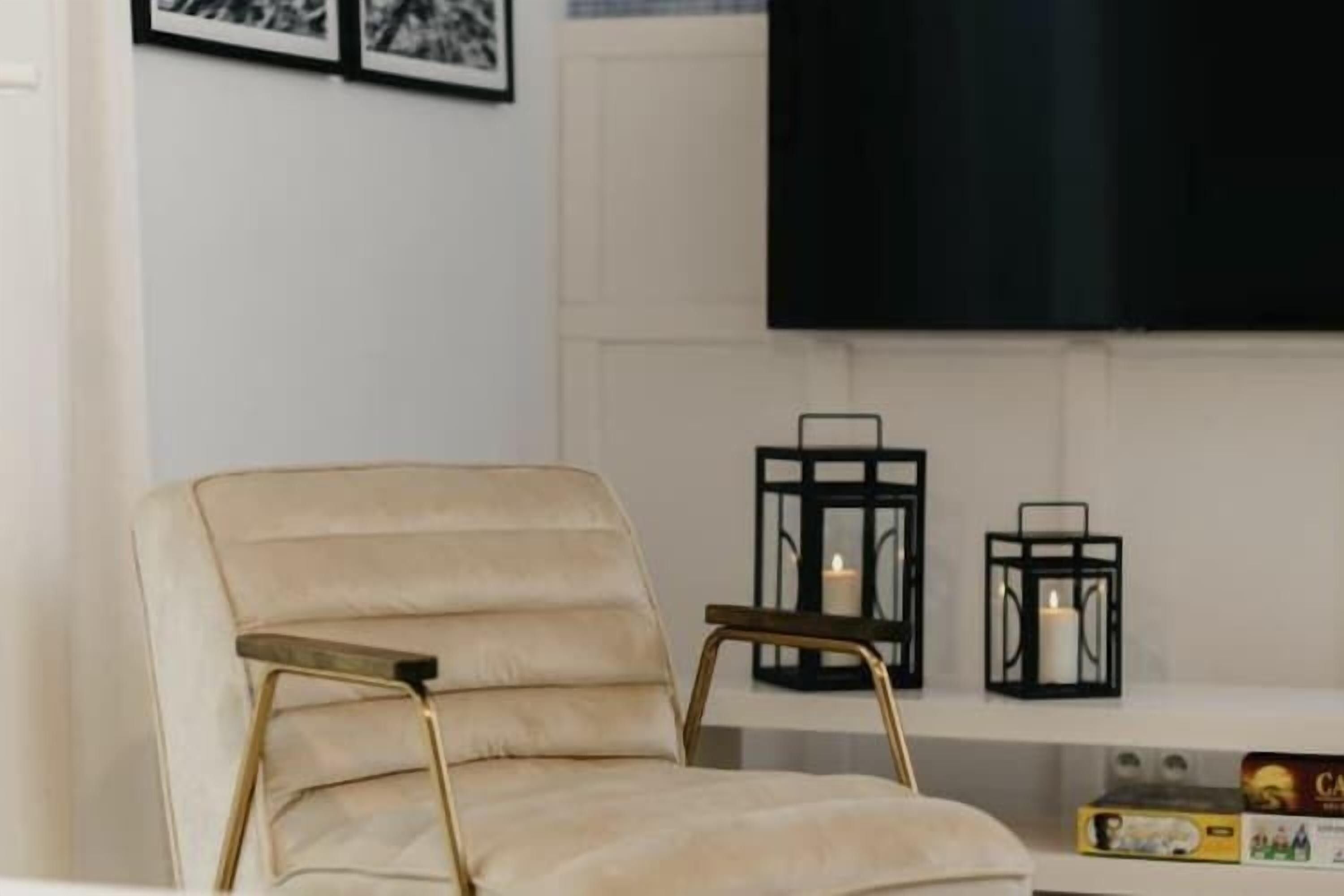 Un fauteuil beige aux pieds dorés, un téléviseur noir accroché au mur et deux chandeliers noirs avec des bougies.