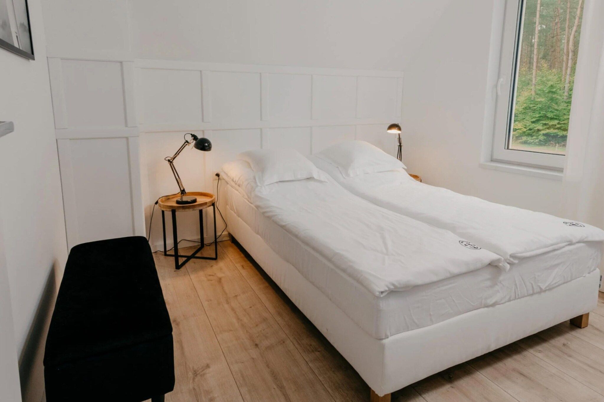 Een kamer met een enkel bed, een houten vloer, witte muren, een raam met uitzicht op bomen en twee bedlampjes.
