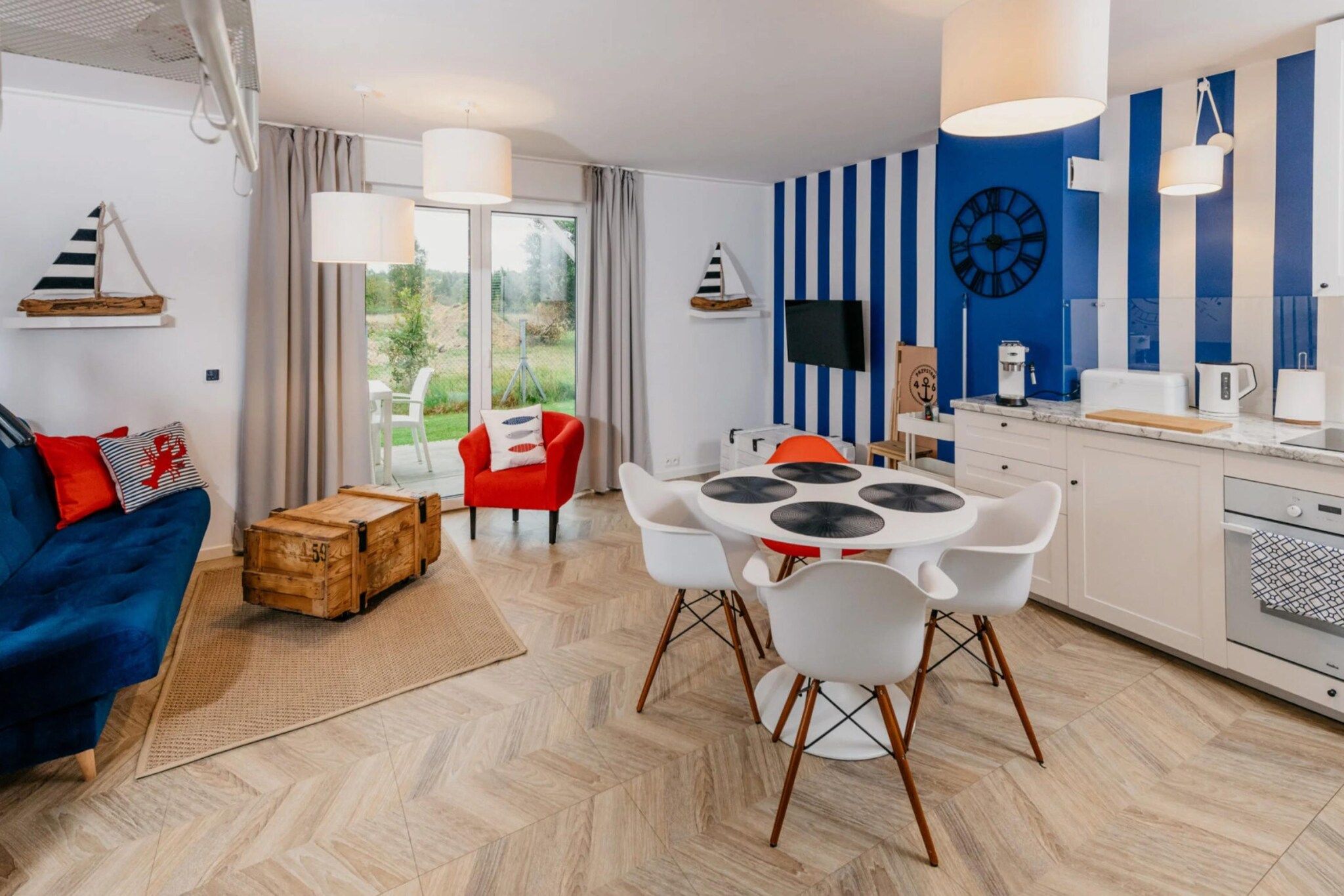 A modern living room with a blue sofa, a red chair, a dining table with chairs, and a kitchen area with a TV and appliances.