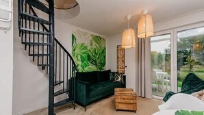 Ein modernes Interieur mit einer Wendeltreppe, einem grünen Sofa, einer Holzkiste und einem großen Fenster mit Blick auf Grünflächen.
