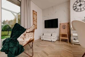 A modern living room with a large window, a flat-screen TV mounted on the wall, a wooden chair, and a white storage unit. - Large holiday home, jacuzzi, sauna (Kopalino)