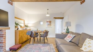 Een gezellige woonkamer met een bank, een eettafel met stoelen, een open haard en een keukenhoek met een houten kast en een aan de muur bevestigde televisie.