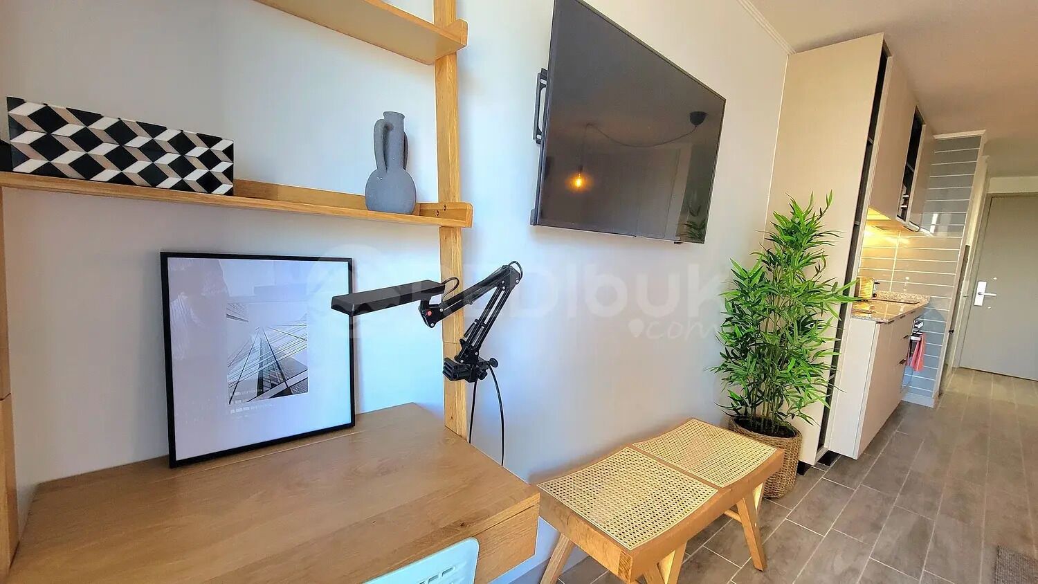 A modern room with a desk, a mounted TV, a framed picture, a camera on a tripod, a small bench, and a potted plant.