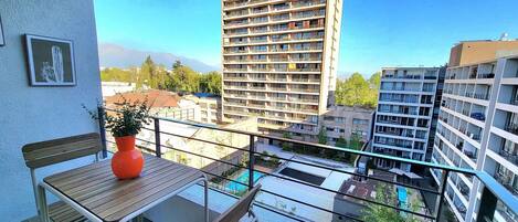 A balcony with a wooden table, a potted plant, and a view of a high-rise building and other residential buildings.