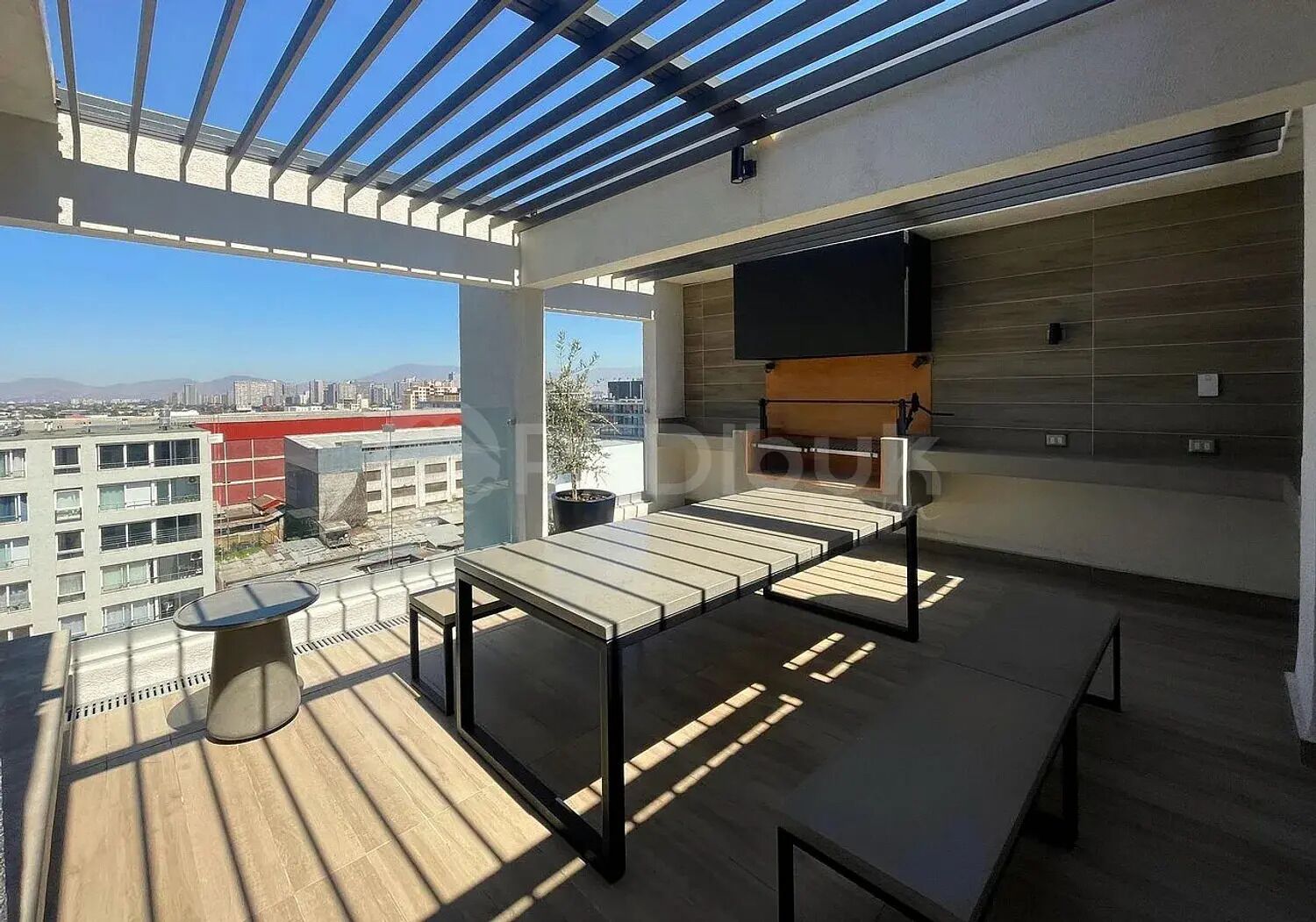 A rooftop terrace with a dining table, chairs, a small round table, a potted plant, and a flat-screen TV mounted on the wall.