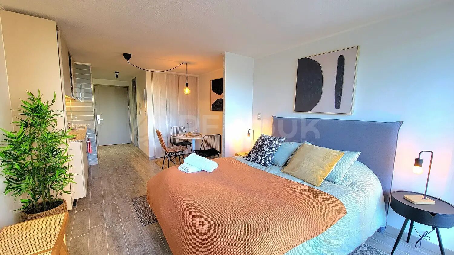 A hotel room with a bed, a bedside table, a lamp, a potted plant, and a view of the hallway.