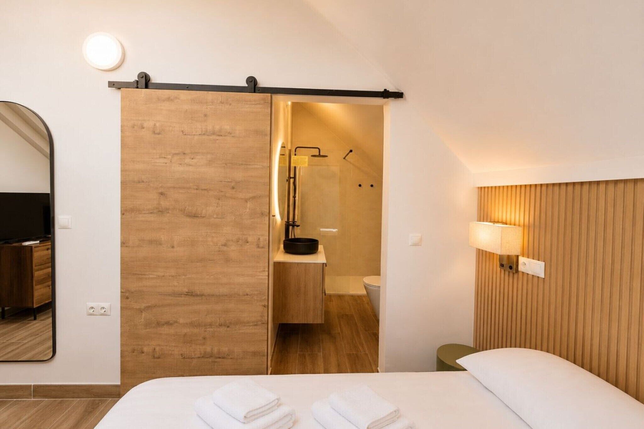 Et hotelværelse med en seng, en skydedør til et badeværelse, et spejl og en lampe monteret på væggen.