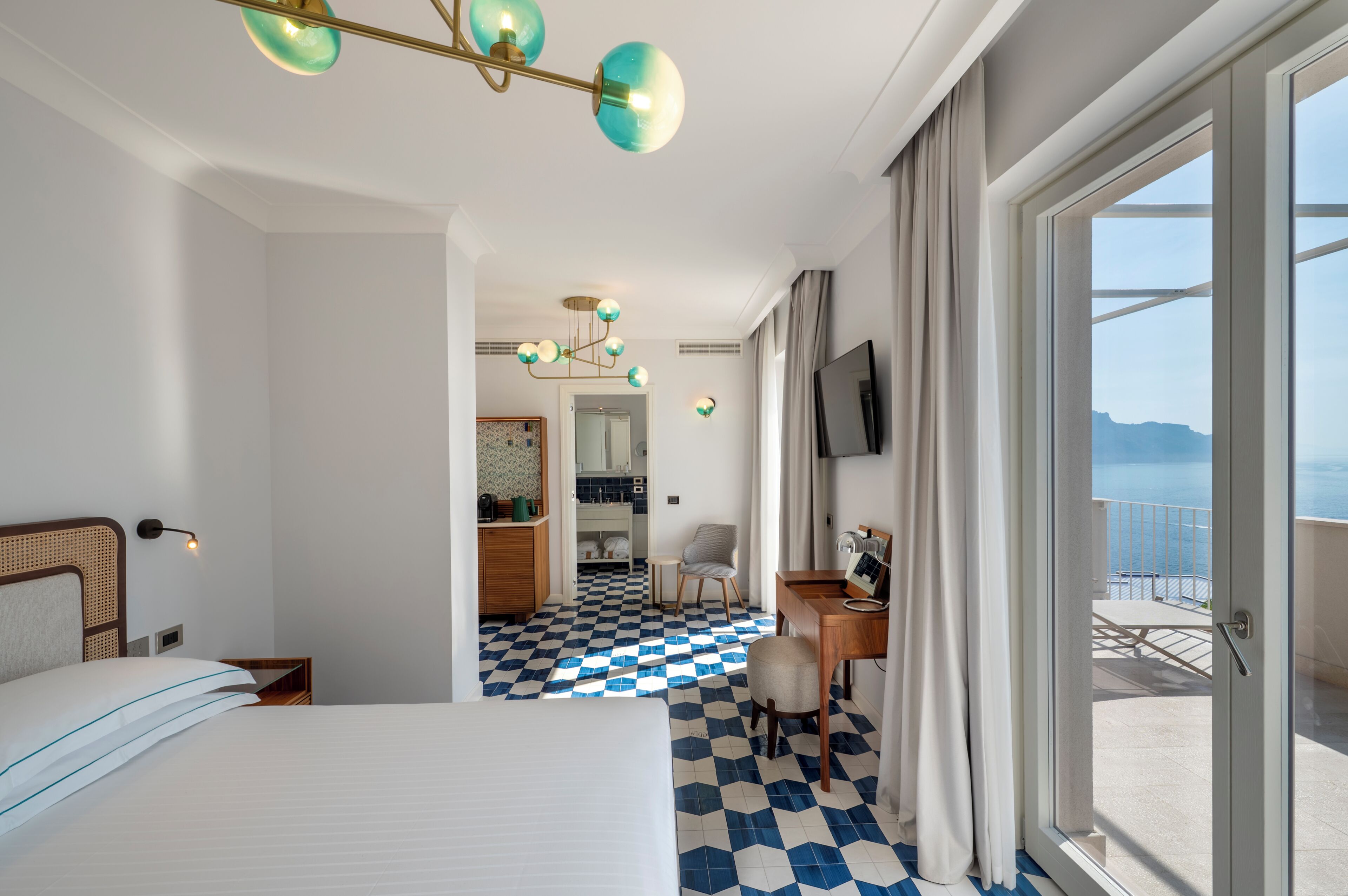 Habitación de hotel con piso a cuadros, una cama, un escritorio con silla, televisión y un balcón con vistas.