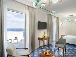 Habitación de hotel con una ventana grande, una cama, un televisor, una mesita con un frutero y una silla.