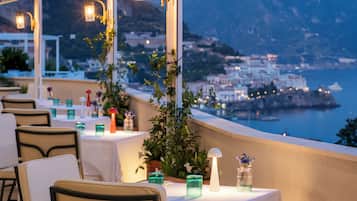 An outdoor dining area with tables and chairs, overlooking a coastal town and the sea.