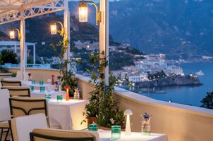 Un área de comedor al aire libre con mesas y sillas, con vistas a un pueblo costero y al mar.