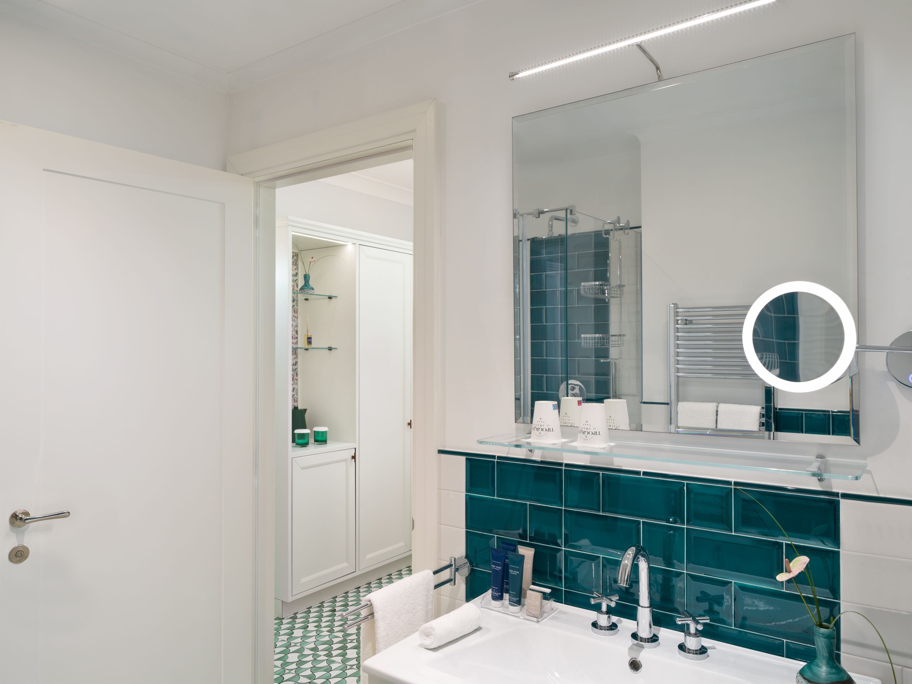 A modern bathroom with a glass shower enclosure, a large mirror with a round light, and a white sink with a chrome faucet.