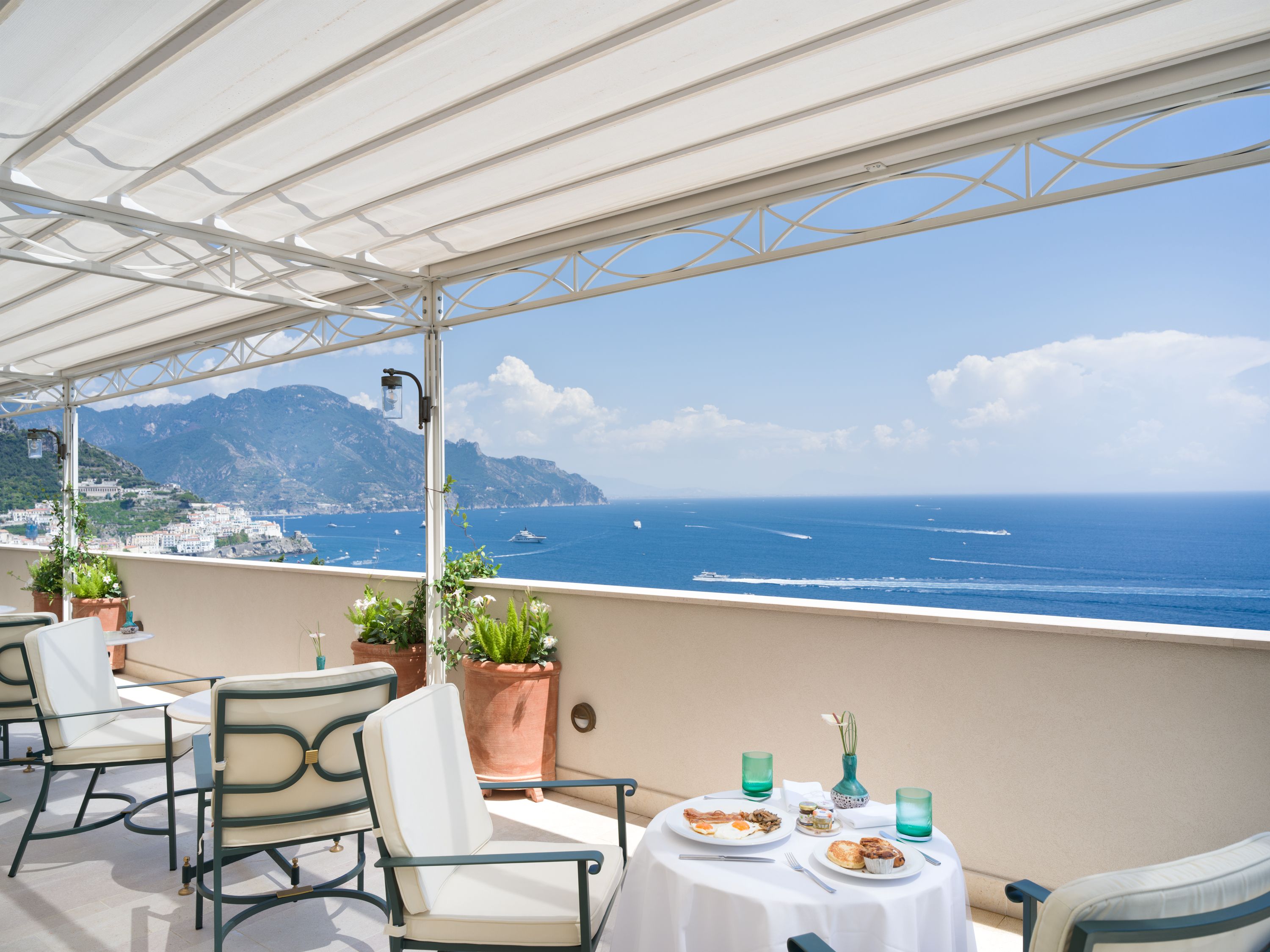 A terrace with outdoor seating, a table set for breakfast, and a view of the sea and mountains.