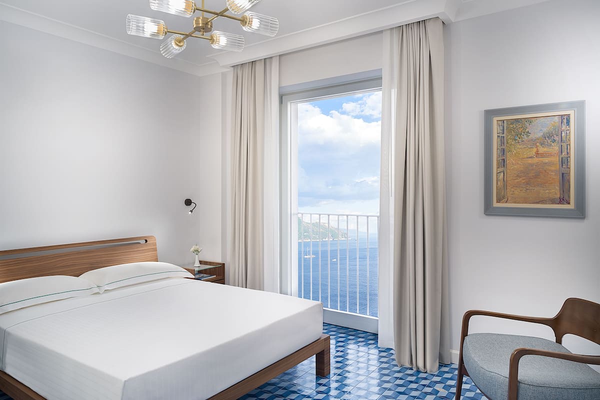 A hotel room with a bed, a chair, a painting, a window with curtains, and a ceiling light fixture.