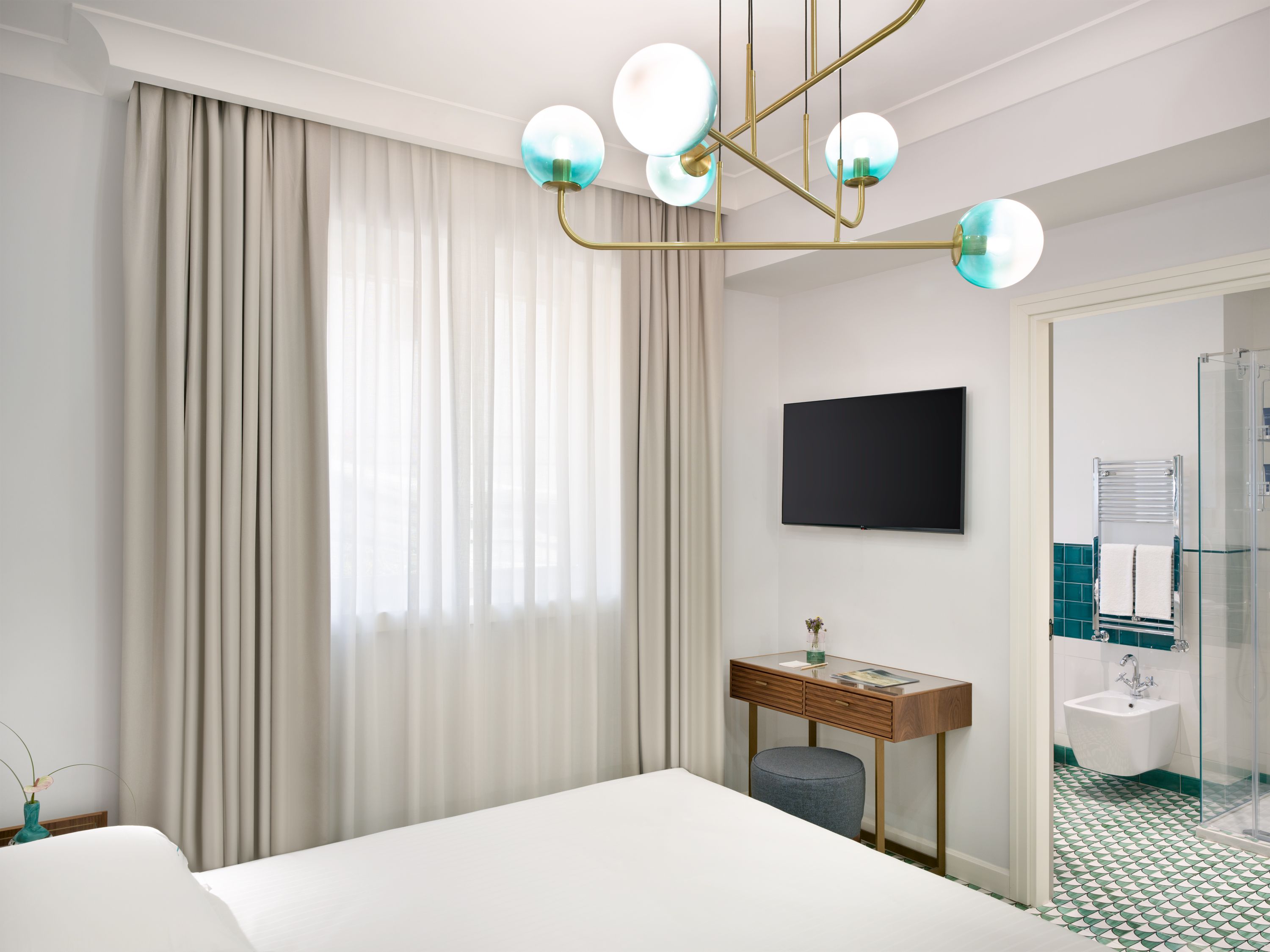 Habitación de hotel con cama, escritorio, televisor, baño con ducha y un gran ventanal con cortinas.