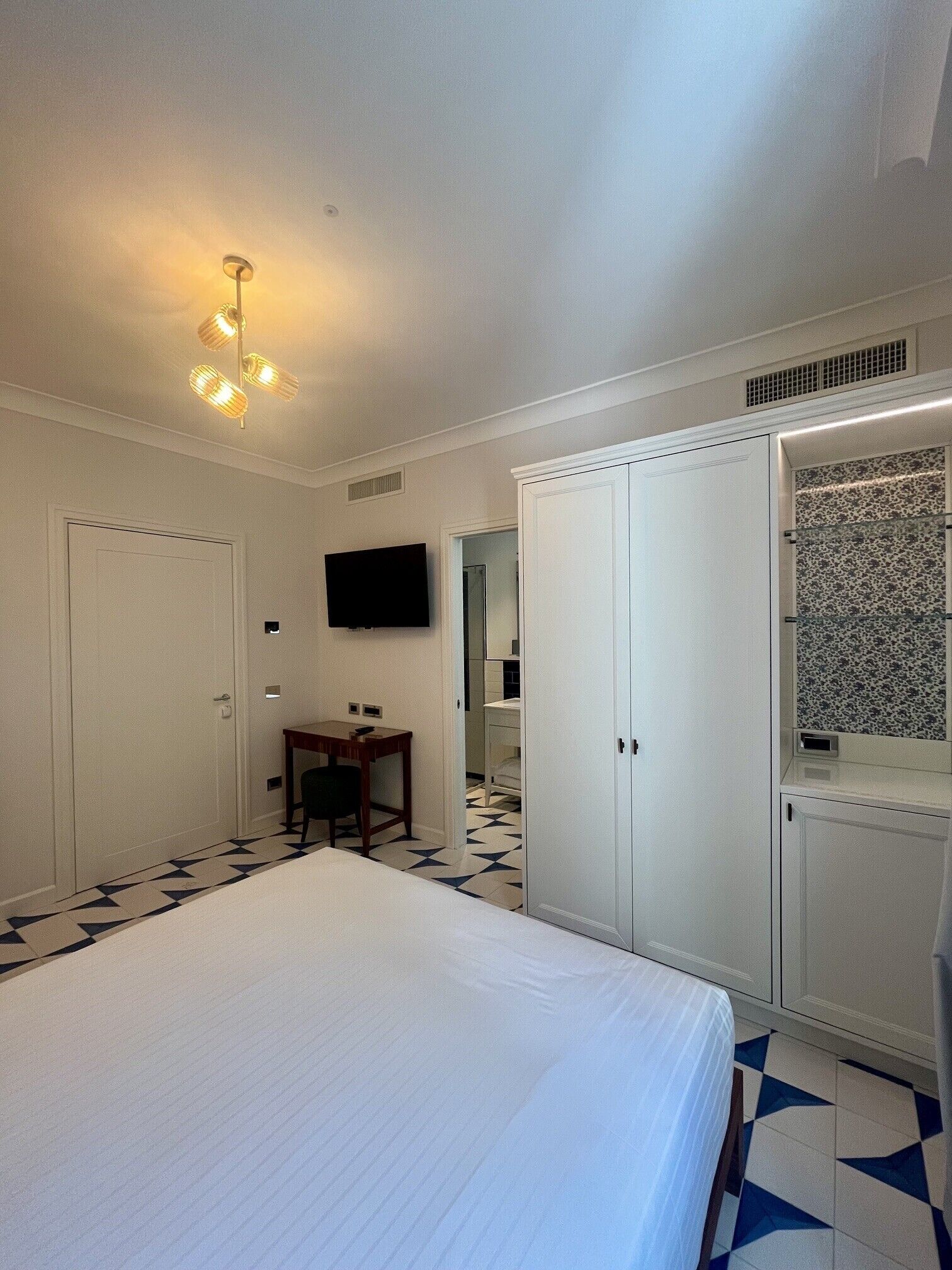 A hotel room with a bed, a desk, a chair, a TV, a wardrobe, and a patterned floor.