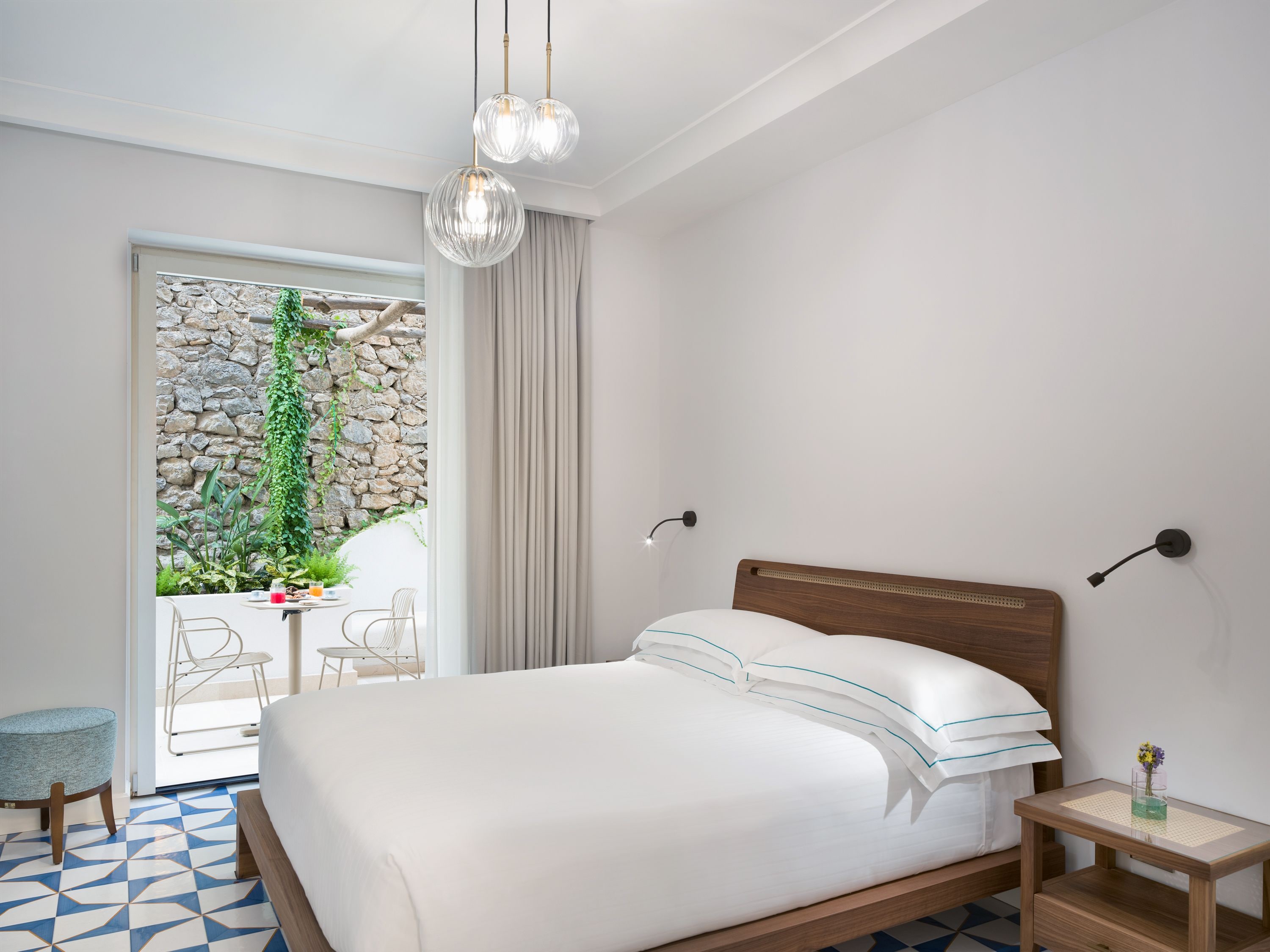A bedroom with a bed, bedside table, and a view of a stone wall and plants.