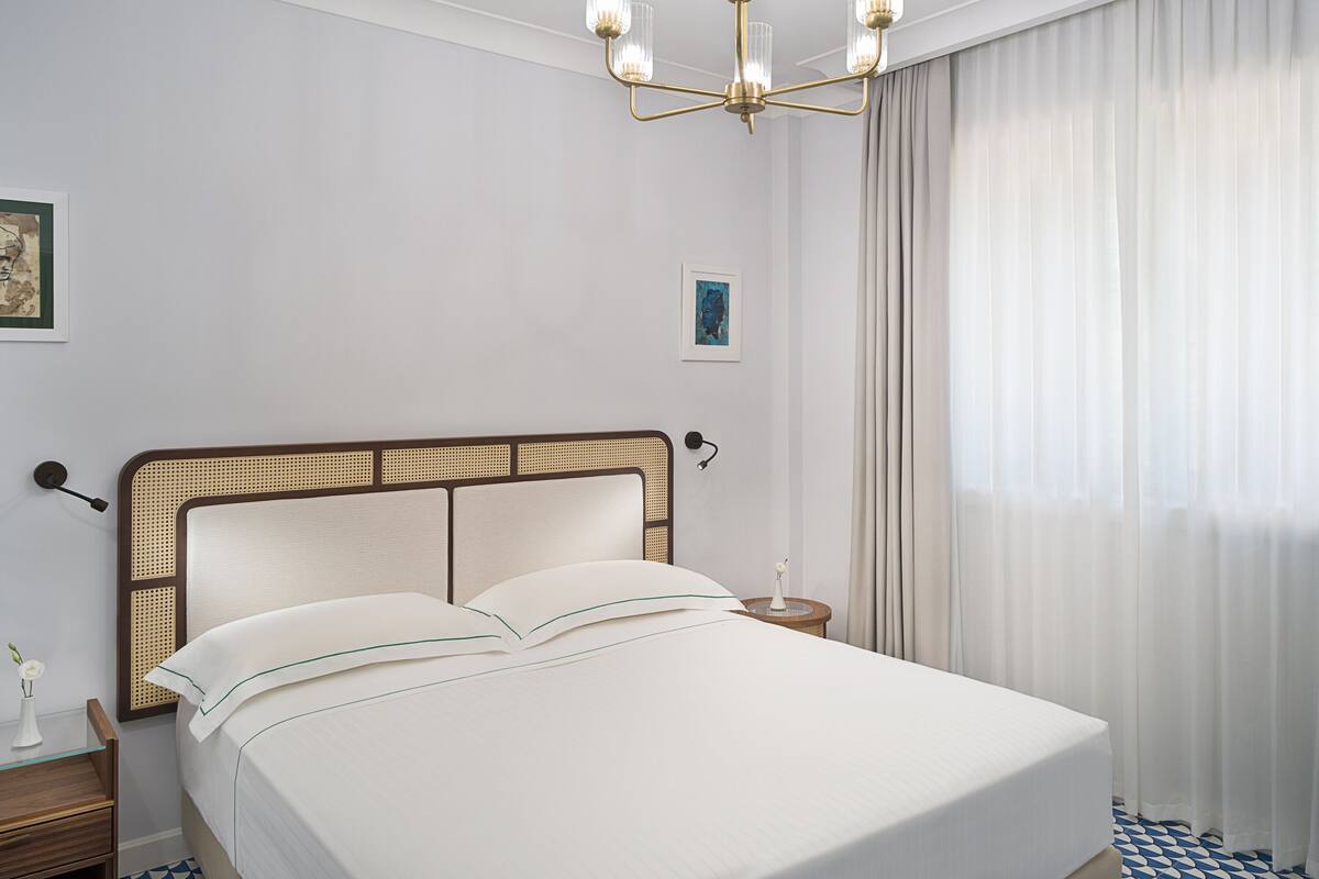 A hotel room with a bed, a chandelier, a nightstand, a vase with a flower, and a picture on the wall.
