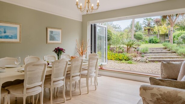 A dining area with a table set for six, surrounded by white chairs, a chandelier, and a view of a garden through large windows. - 5 Bed in Seaview (oc-n33058) (Seaview)