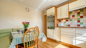 A compact kitchen with a dining area, featuring a checkered tablecloth, wooden chairs, a clock, and a tiled backsplash. - 4 Bed in Harlyn Bay (oc-t35030) (Harlyn Bay)