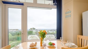 Een eetgedeelte met een tafel gedekt voor twee personen, een vaas met bloemen en uitzicht op zee door grote ramen.