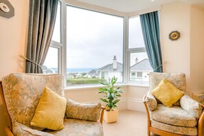 Ein Zimmer mit zwei Sesseln, einer Topfpflanze und Blick auf Häuser durch große Fenster.