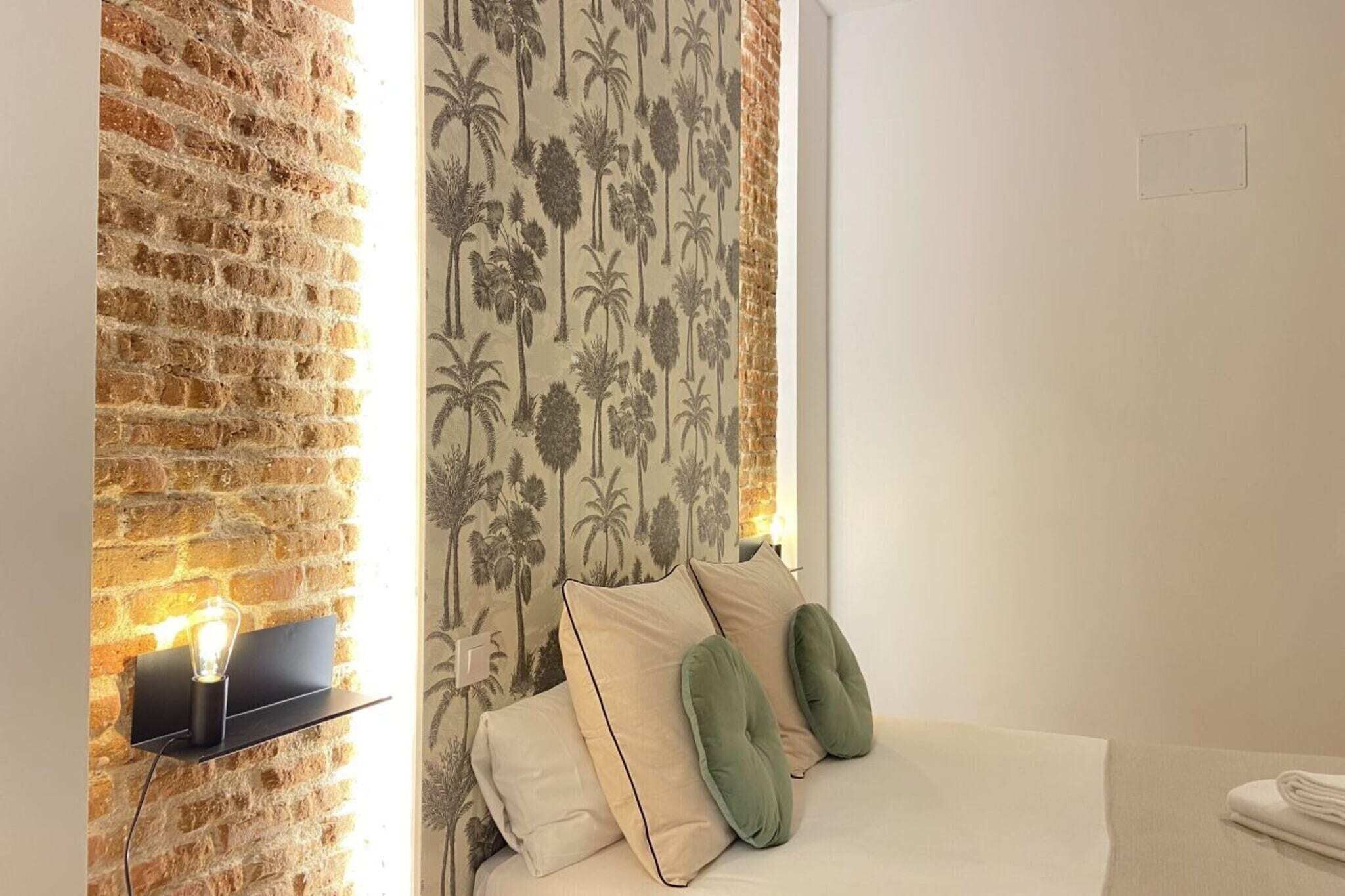 Una habitación con pared de ladrillo, una cama con almohadas, una lámpara de pared y un papel tapiz estampado.