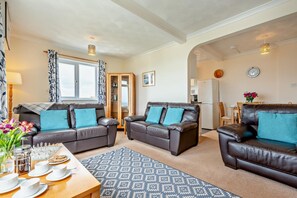 A living room with two leather sofas, a patterned rug, and a dining area with a table set for a meal. - 2 Bed in Harlyn Bay (oc-s35034) (Harlyn Bay)