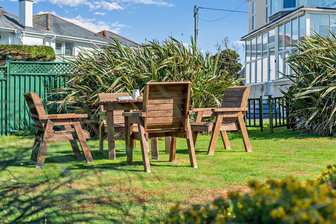Zona relax esterna in legno con tavolo e sedie, immersa nel verde e con un edificio bianco sullo sfondo.