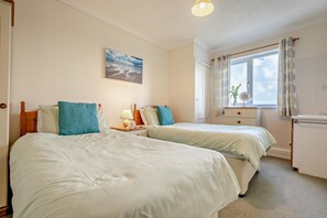 A room with two beds, a bedside table with a lamp, a window with curtains, and a painting on the wall. - 2 Bed in Harlyn Bay (oc-s35034) (Harlyn Bay)