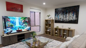 Ett vardagsrum med en stor tv-skärm, ett soffbord i glas, en beige soffa, hyllor i trä och en stor, abstrakt målning.