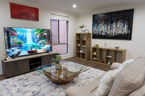 A living room with a large flat-screen TV, a glass coffee table, a beige sofa, wooden shelves, and a large abstract painting. - 47A Westlake Road Morley is a Perfect 4-bedroom house with AC in gorgeous Morley (Morley)