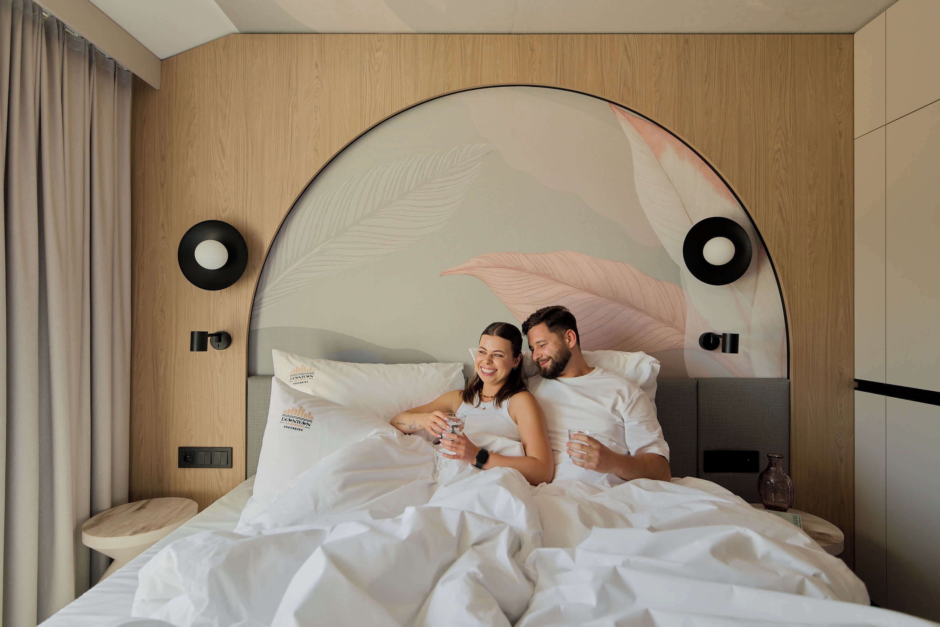 A bed with white bedding, a wooden headboard, and a wall-mounted mirror.