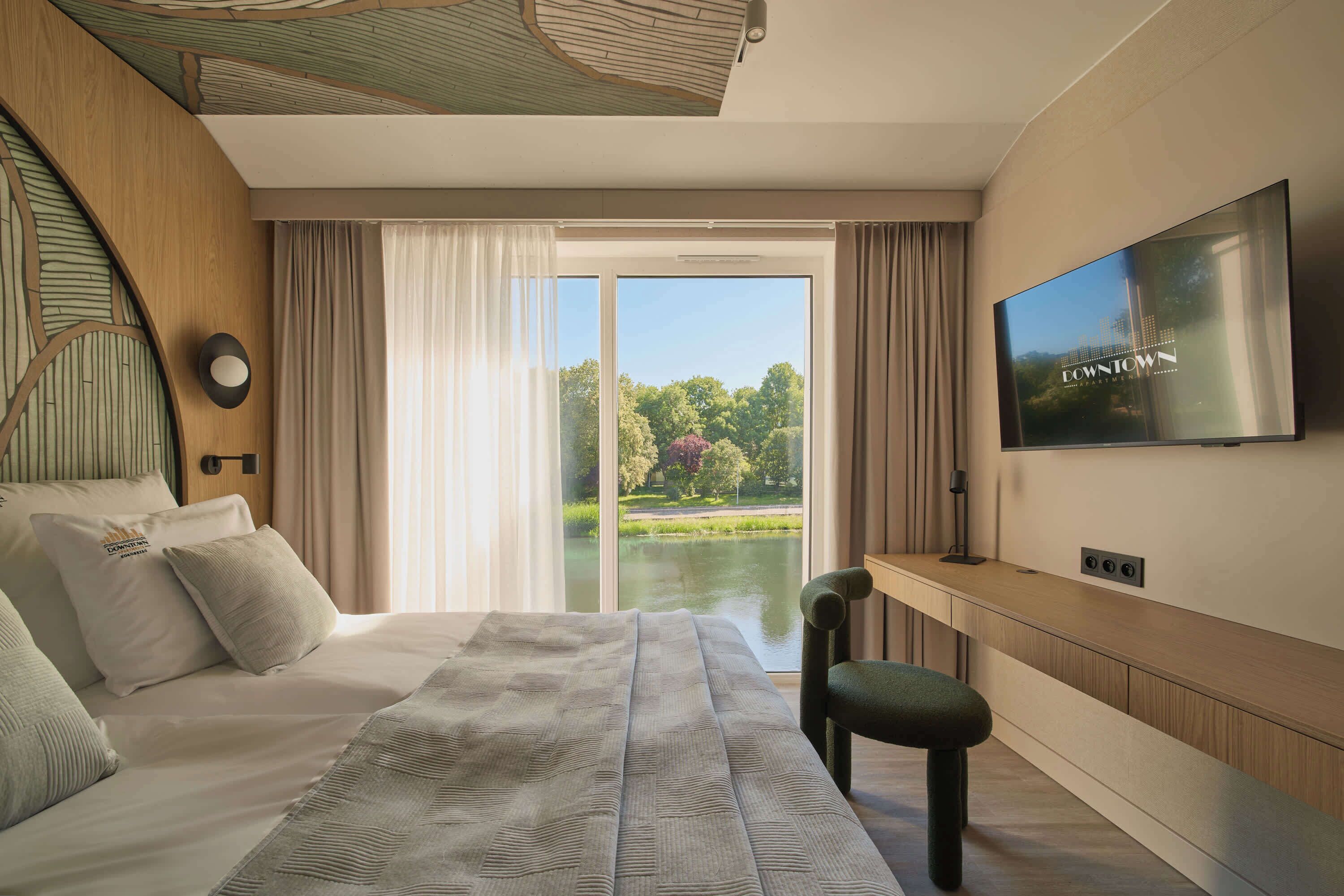 A bedroom with a large bed, a TV mounted on the wall, a desk, and a view of a park through the window.