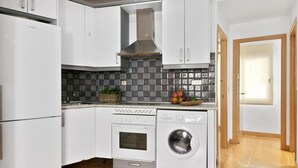 Una cocina compacta con muebles blancos, horno integrado, lavadora y salpicadero de azulejos.