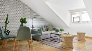 Un dormitorio tipo loft moderno con techo inclinado, papel tapiz estampado, un sofá gris, dos sillones, una mesita y una ventana.