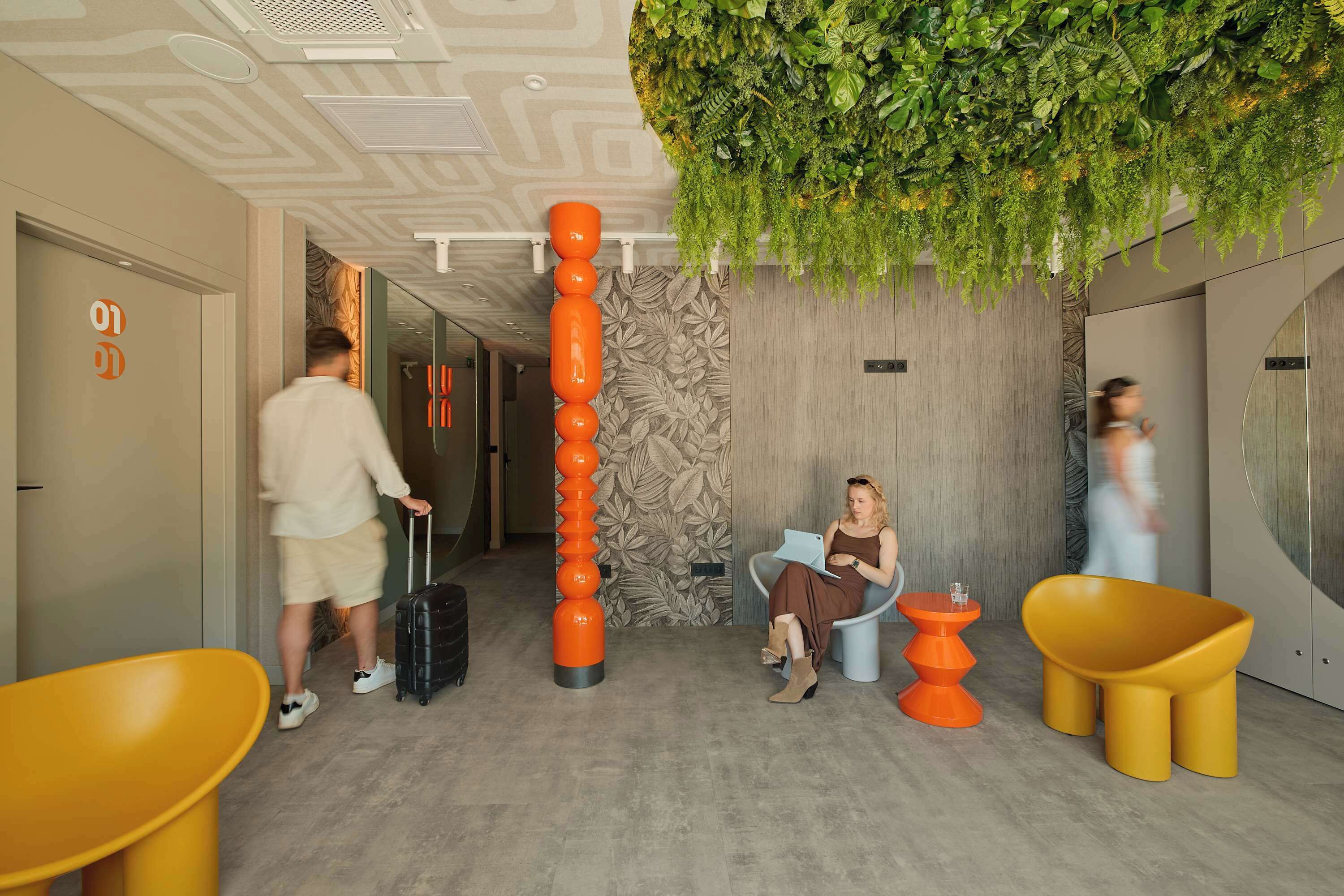 A hotel lobby with a modern design, including a vertical orange column, a seating area with yellow chairs, and a green wall with hanging plants.