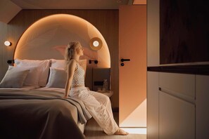 A modern bedroom with a curved headboard, a bedside table with a glass container, and a view of the outside. - Persante Sunrise Collection 11 | Riverview | AC (Kołobrzeg)