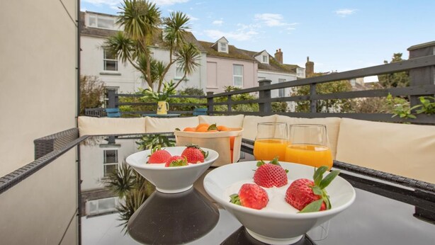 A balcony with two bowls of strawberries and glasses of orange juice, a wicker chair, and a view of houses and palm trees. - 4 Bed in Appledore (oc-sunni) (Appledore)