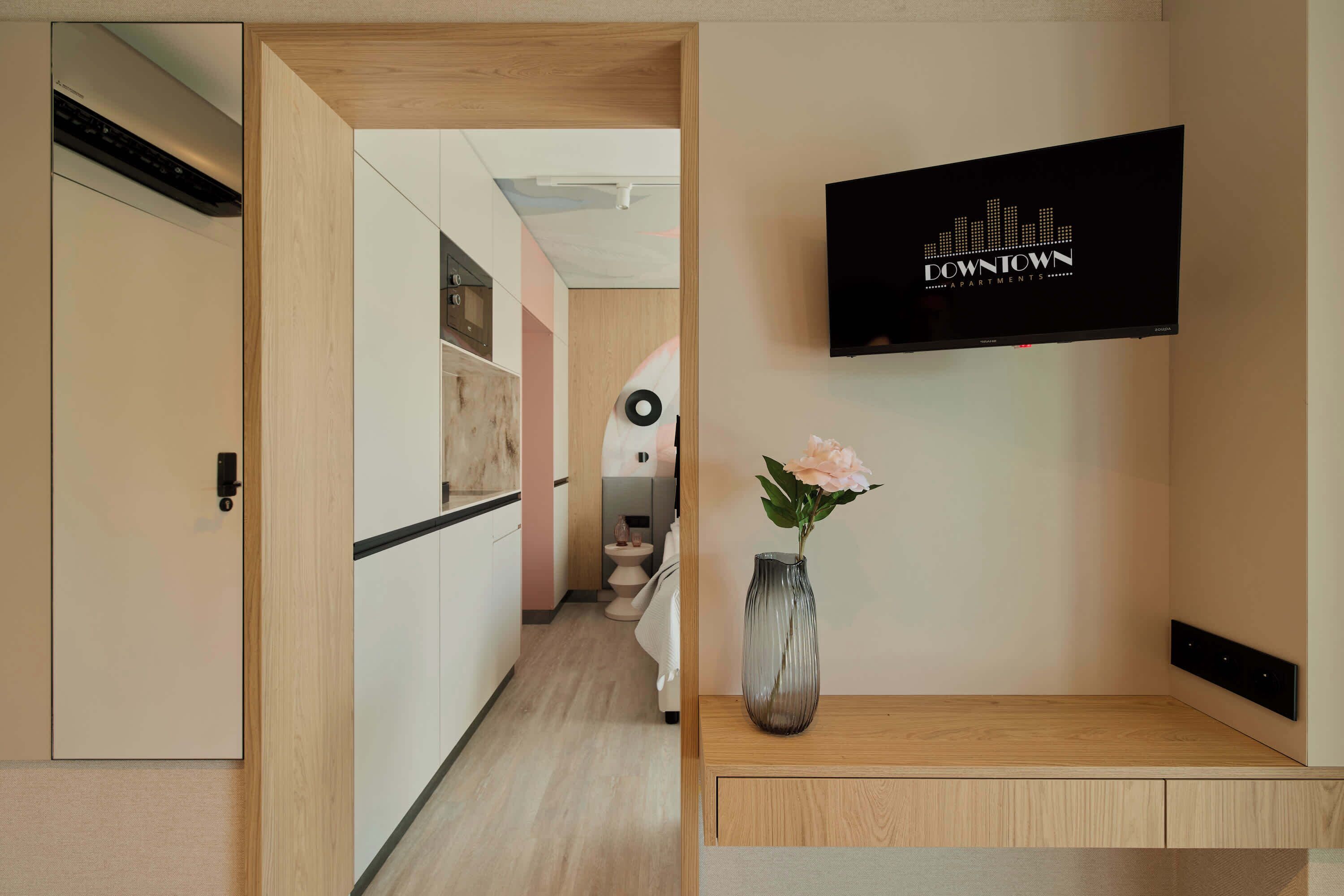 Un couloir avec une porte en bois, un téléviseur fixé au mur qui affiche le texte « APPARTEMENTS DU CENTRE-VILLE », et un vase avec une fleur posé sur une console en bois.
