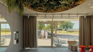 Um saguão de hotel moderno com porta de vidro que dá para o exterior, bebedouro e área de estar com cadeiras laranja.
