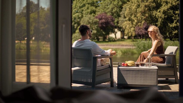 Um casal sentado em móveis de pátio ao ar livre, de frente para um corpo d'água, com uma mesa de vime entre eles, com bebidas e petiscos.