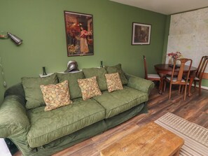 Een groene bank met bloemkussens, een houten salontafel, een eettafel met stoelen en twee afbeeldingen in lijsten aan de muur.