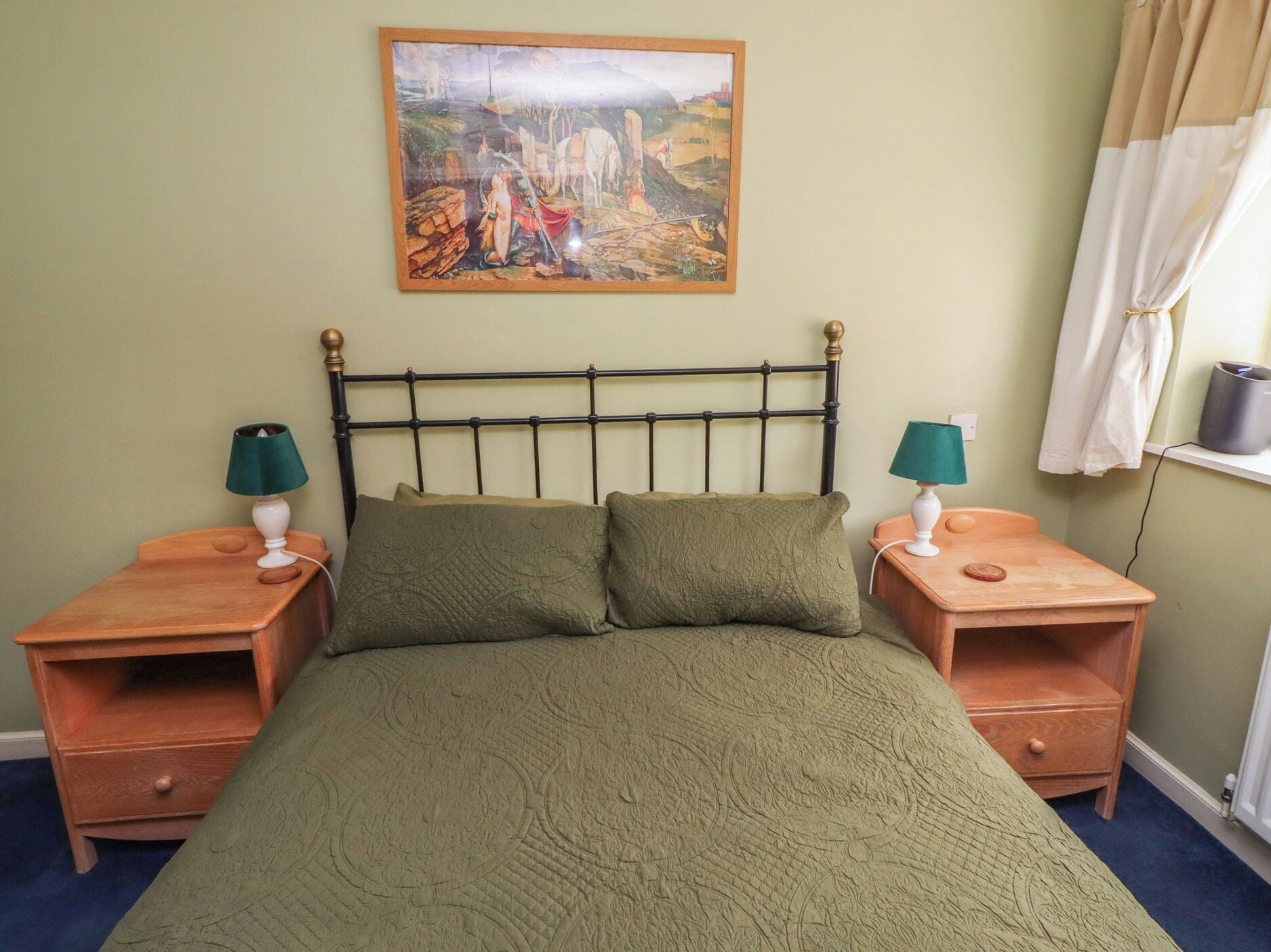A bed with a green quilt, two bedside tables with lamps, a painting on the wall, and a window with curtains.