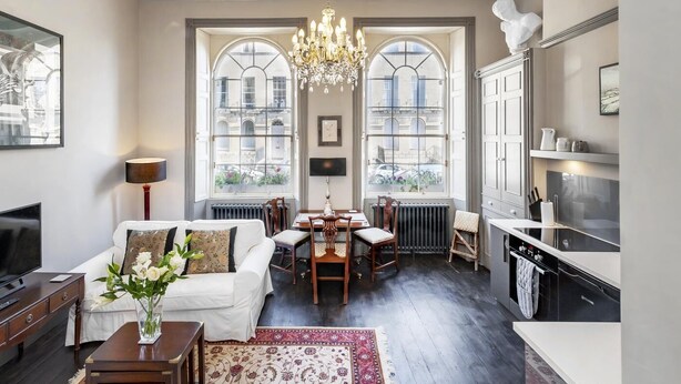 A spacious living room with a white sofa, dining table, chandelier, and large windows. - 1 Bed in Bath (oc-bhr33) (Bath)