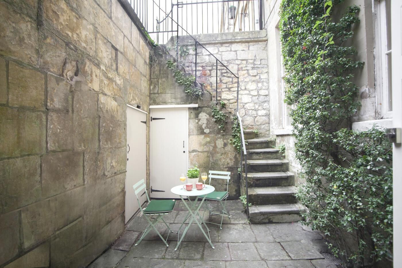 A stone-walled courtyard with a small table and chairs, a white door, and a staircase.