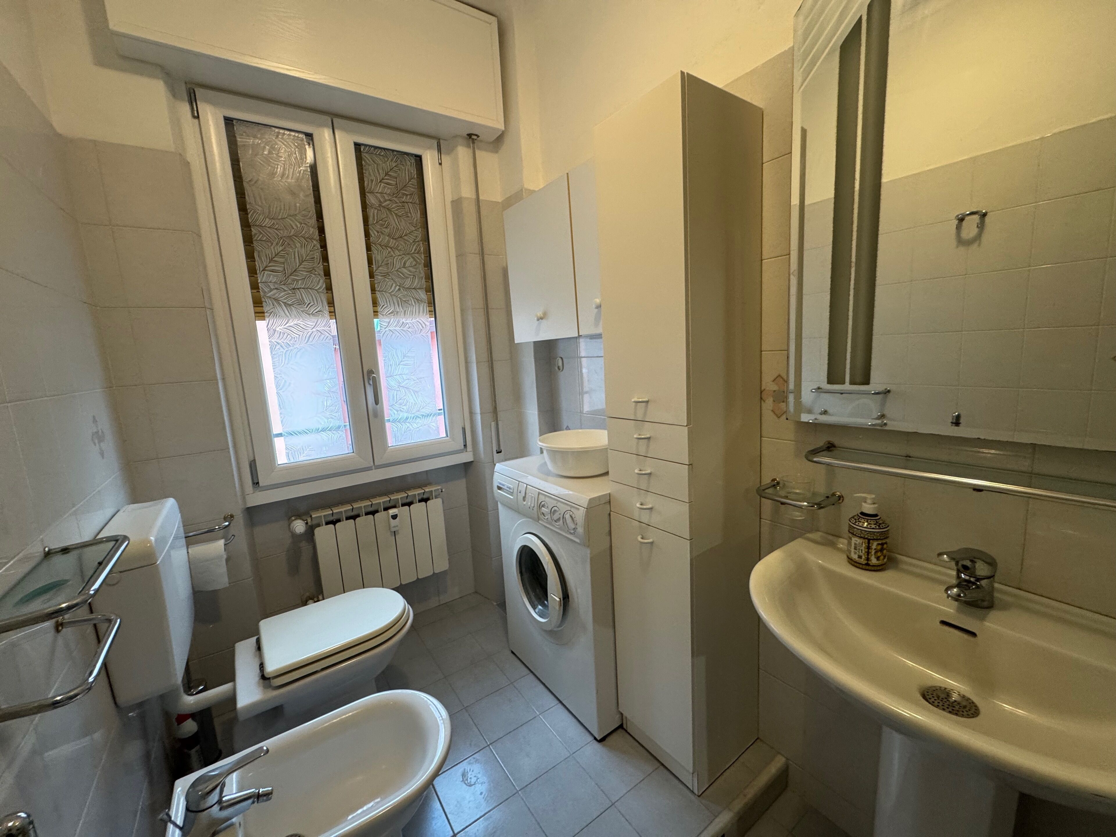 A bathroom with a toilet, washing machine, sink, and a window with frosted glass.