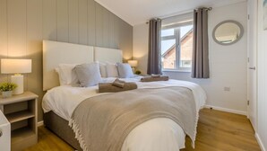 Ett sovrum med en säng, sängbord, en lampa, ett fönster med gardiner, en spegel och utsikt över en byggnad utanför.