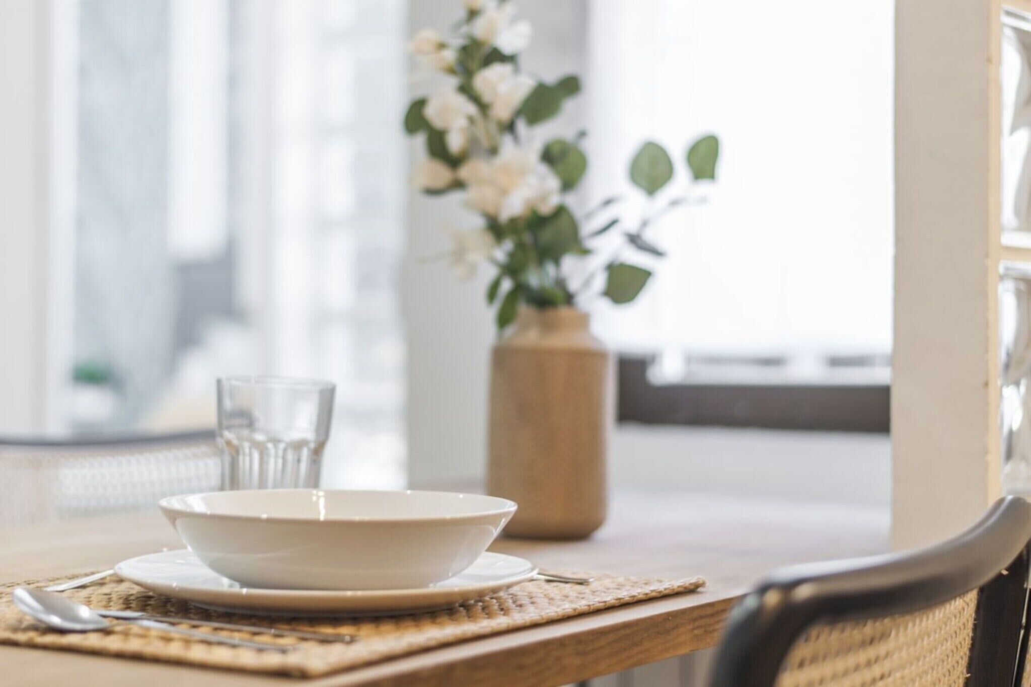 A dining table with a bowl and glass, a woven placemat, and a vase with flowers.